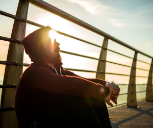 A man in a hood sitting down, resting against a metal barrier at sunset. 