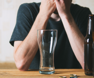 A close up of an empty pint glass in front a male whose face isn't visible. A beer bottle and bottle tops are on the table he rests his elbows on. 