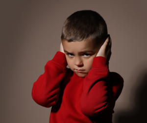 A small boy in a red jumper covering his ears with his hands, looking fearful. 