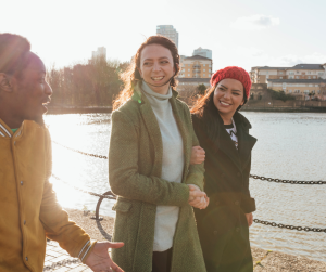 Two ladies link as they walk next to a river chatting with a man. 