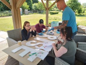 A group of people doing arts and crafts at Dutch Farm. They're gathered around a table, decorating large letters reading R I S E