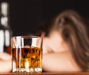 An out of focus female with her head in her hands behind a glass of a dark spirit such as Brandy or Whiskey. 