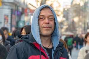 A middle aged man wearing a hoody on a busy street smiles at the camera