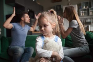 A young girl cuddling a teddy as her parents argue in the background. 