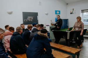 A group meeting at YMCA Together. 8 individuals, back to camera, listen to two speakers sat on a pool table. 